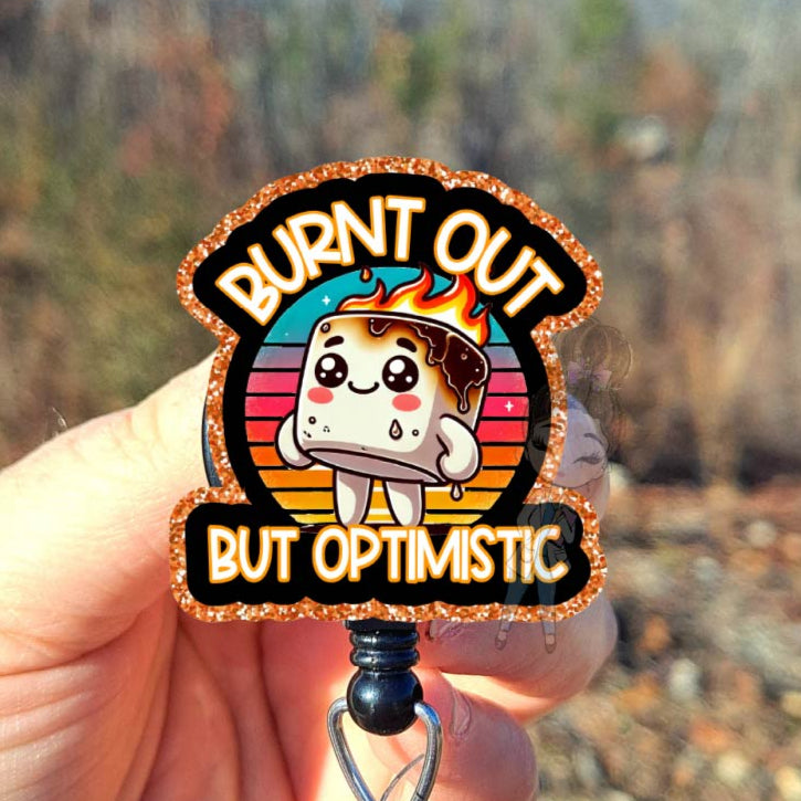 badge reel with cartoon character and text 'Burnt Out But Optimistic' held by a hand outdoors.