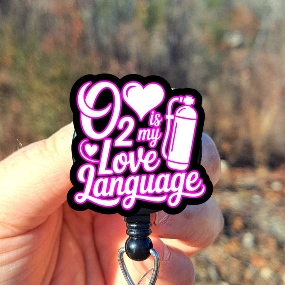 Black badge holder with pink text 'O2 is my Love Language' held by a hand against a blurred natural background.