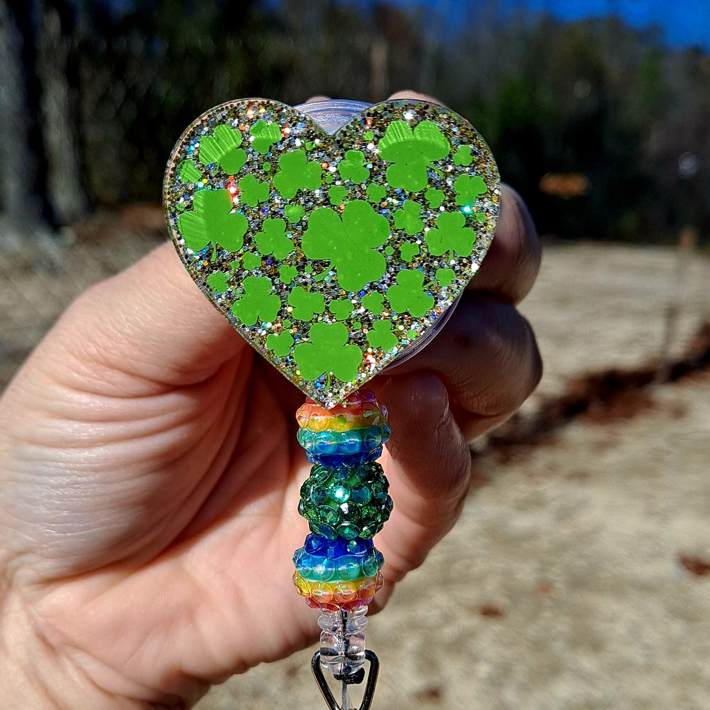 Heart-shaped keychain with green pattern and rainbow base held by a hand outdoors.
