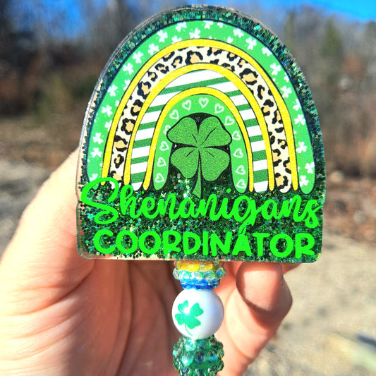 Decorative keychain with rainbow and shamrock design held by a hand outdoors.