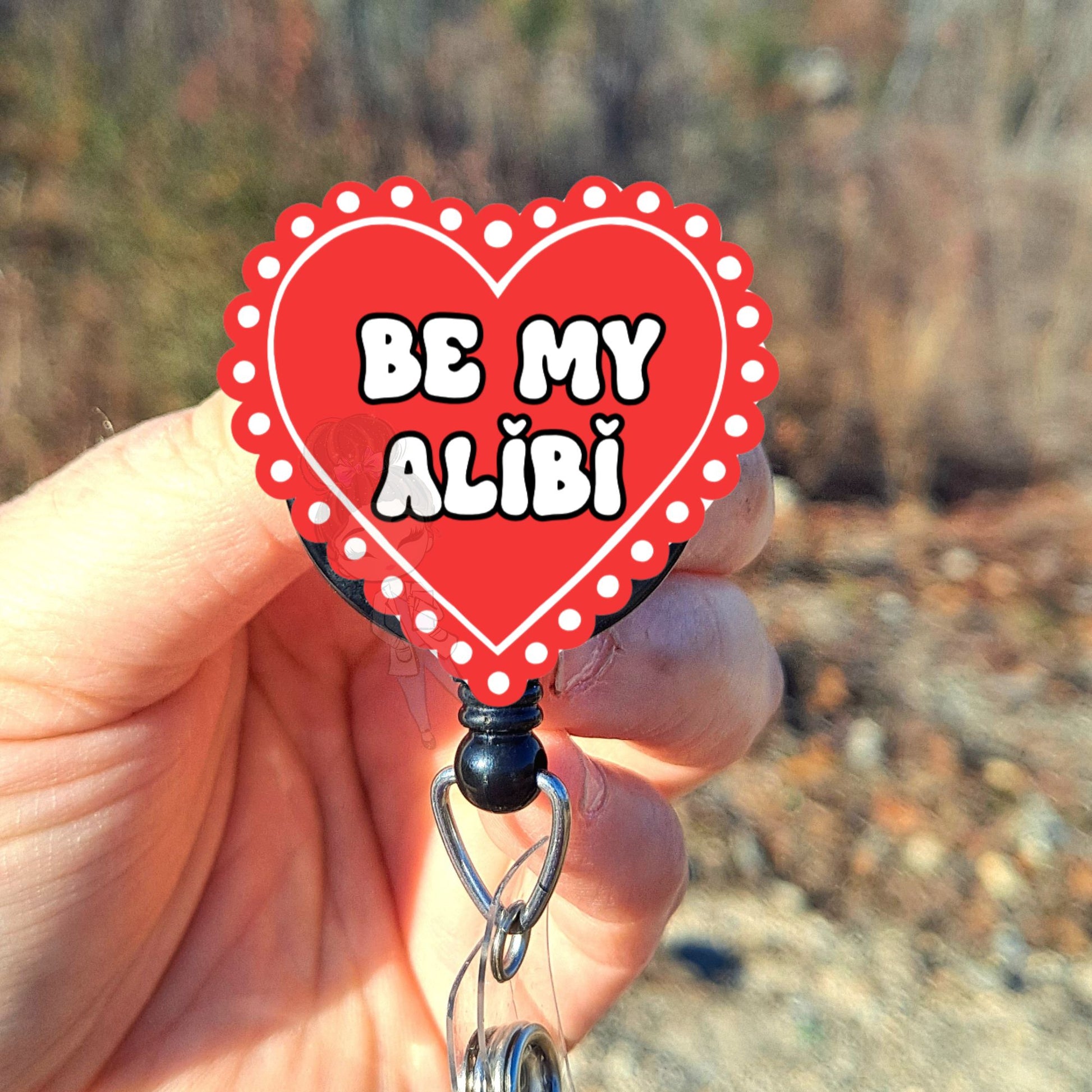 Heart-shaped badge holder with 'Be My Alibi' text held by a hand outdoors.