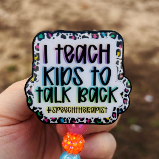 Colorful badge with 'I teach kids to talk back #speechtherapist' text held by a hand against a blurred natural background.