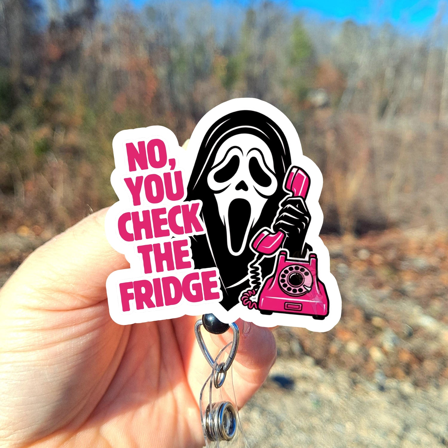 A hand holds a id badge reel featuring a black-and-white Ghost like figure holding a pink rotary phone next to pink text that reads 'NO, YOU CHECK THE FRIDGE.' The background is a blurred outdoor scene with trees and blue sky.
