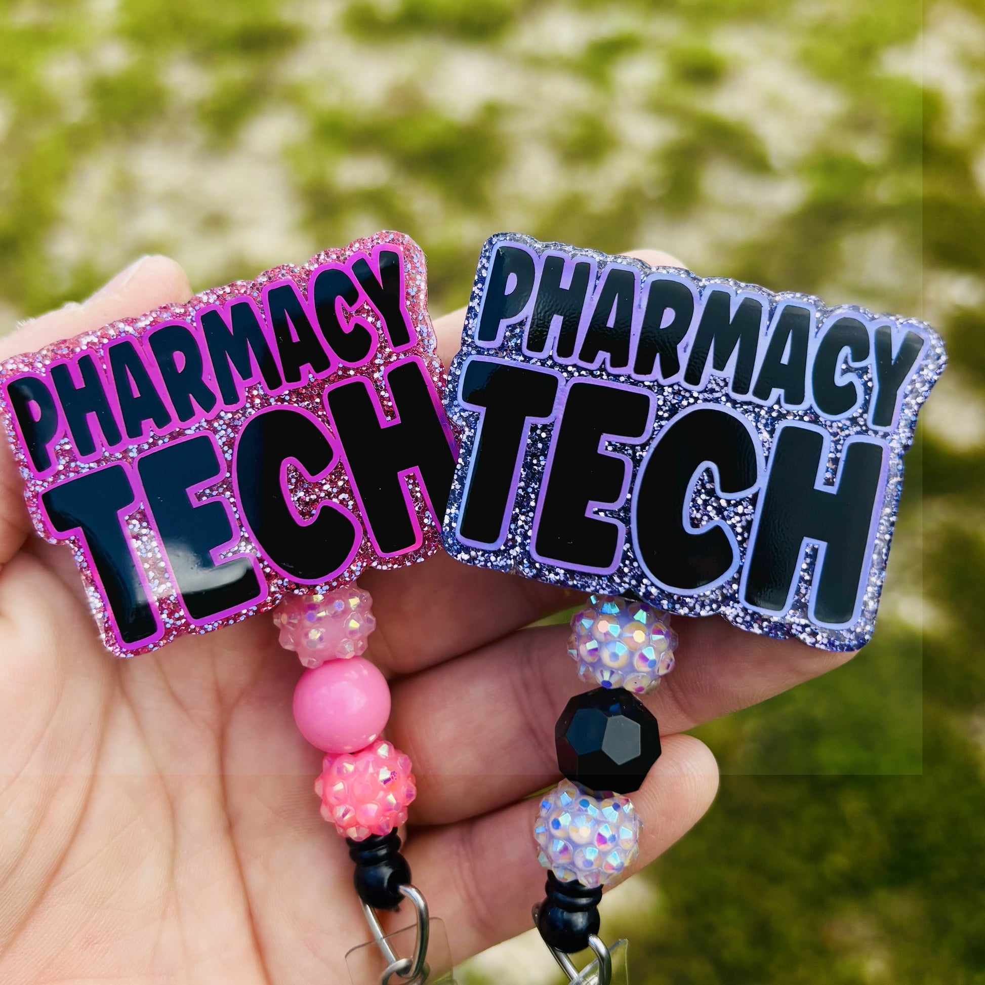 Two 'PHARMACY TECH' badge holders with colorful beads held in a hand against a blurred green background.