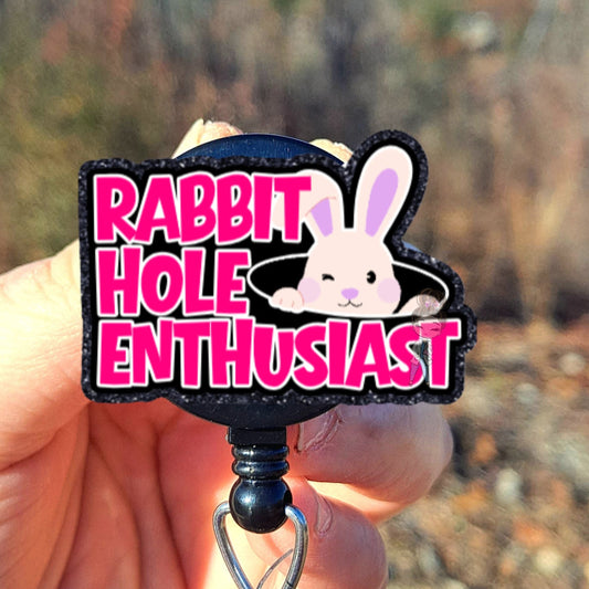 Hand holding a badge reel with 'Rabbit Hole Enthusiast' design against a natural background