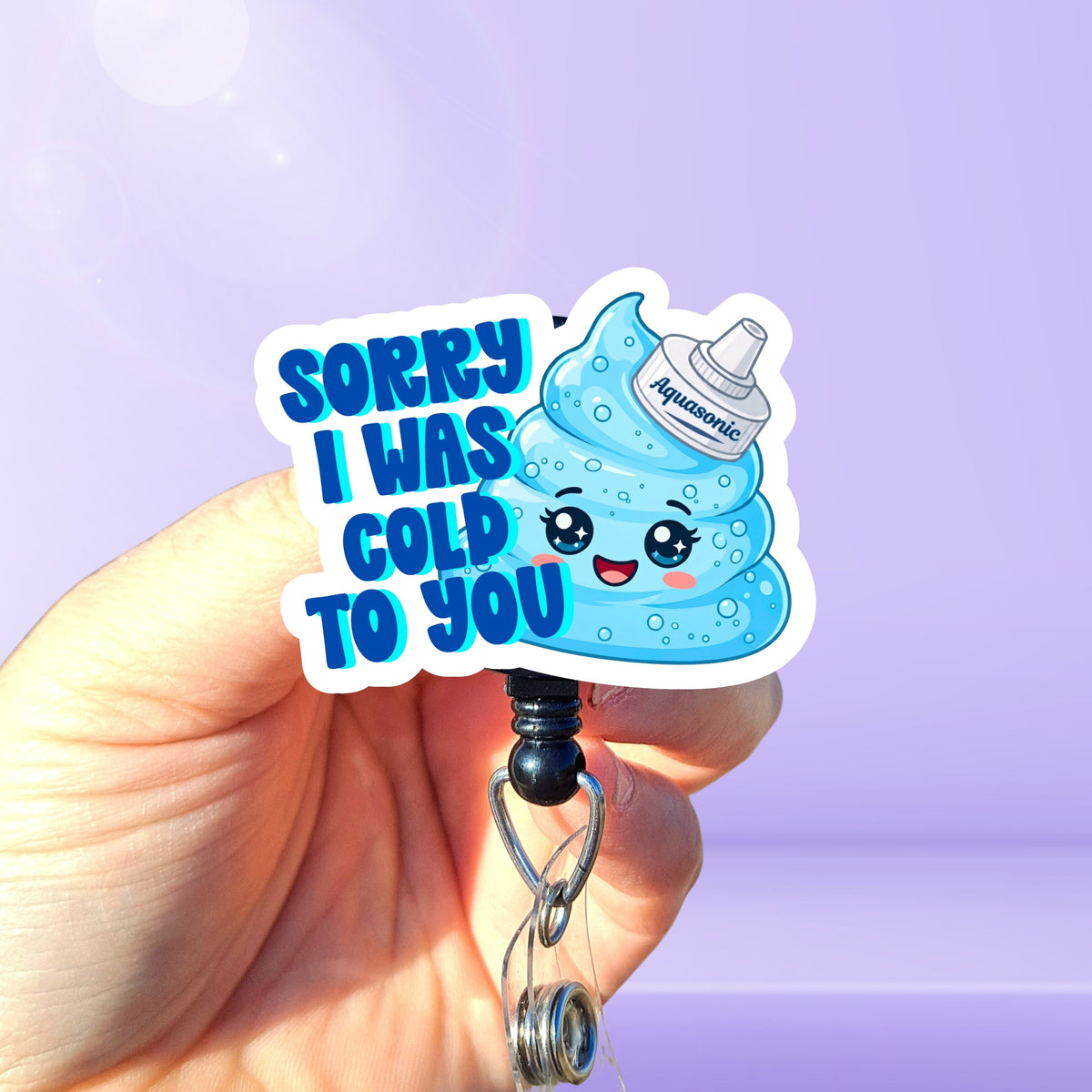 A hand holding a badge reel featuring a cute blue aquasonic gel blob swirl character and the text 'SORRY I 
WAS COLD TO YOU' on a light purple background.
