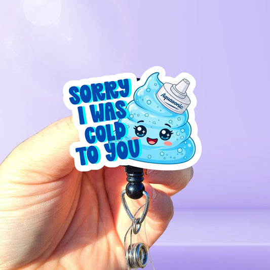 A hand holding a badge reel featuring a cute blue aquasonic gel blob swirl character and the text 'SORRY I 
WAS COLD TO YOU' on a light purple background.